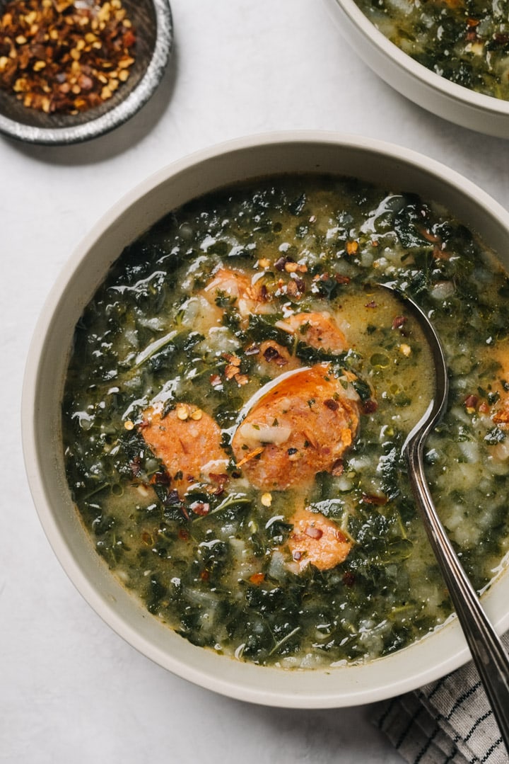 A bowl of caldo verde green soup on a cement background with a small bowl of red pepper flakes to the side.