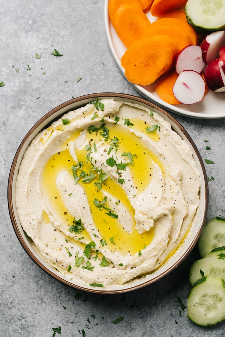 A bowl of cauliflower hummus on a cement background with a plate of raw sliced vegetables.