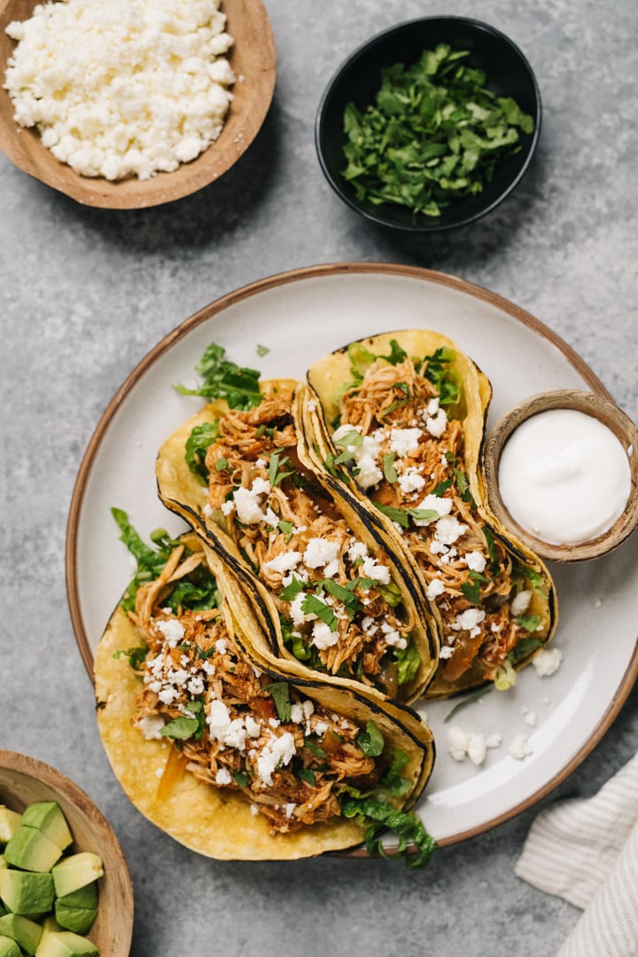 Three chicken tinga tacos in corn tortillas with cilantro and queso fresco on a white plate.