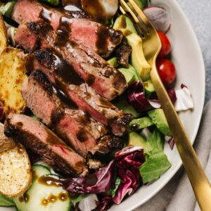Whole30 or keto steak salad in a tan salad bowl with a gold fork - seared sirloin, avocado, salad greens, blue cheese or roasted potatoes, and balsamic dressing.