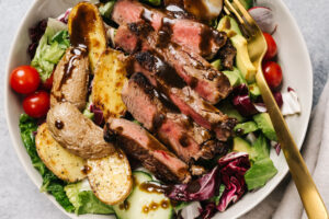 Healthy steak salad with roasted potatoes and avocado in a tan bowl with a gold fork.