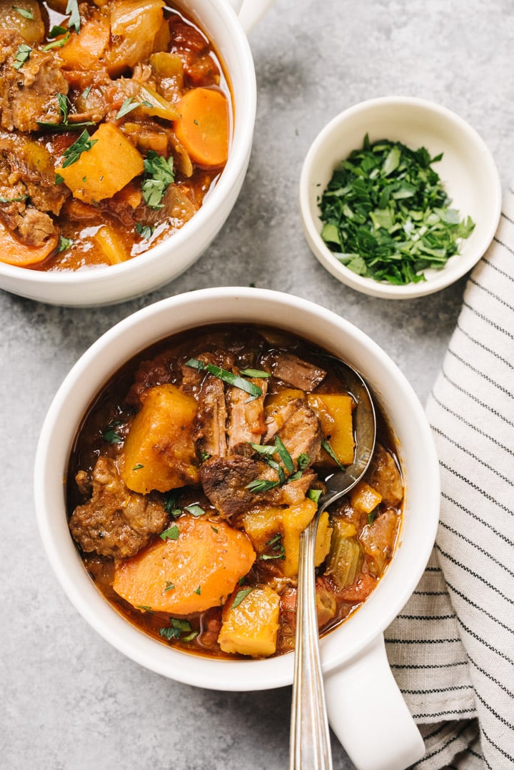 Two bowls of slow cooker whole30 beef stew with butternut squash on a concrete table with a striped linen napkin and small bowl of chopped parsley.