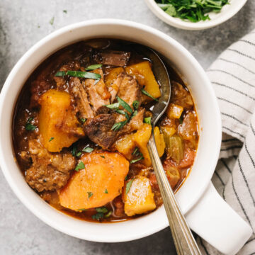 A bowl of slow cooker whole30 beef stew with butternut squash on a concrete table with a striped linen napkin and small bowl of chopped parsley.