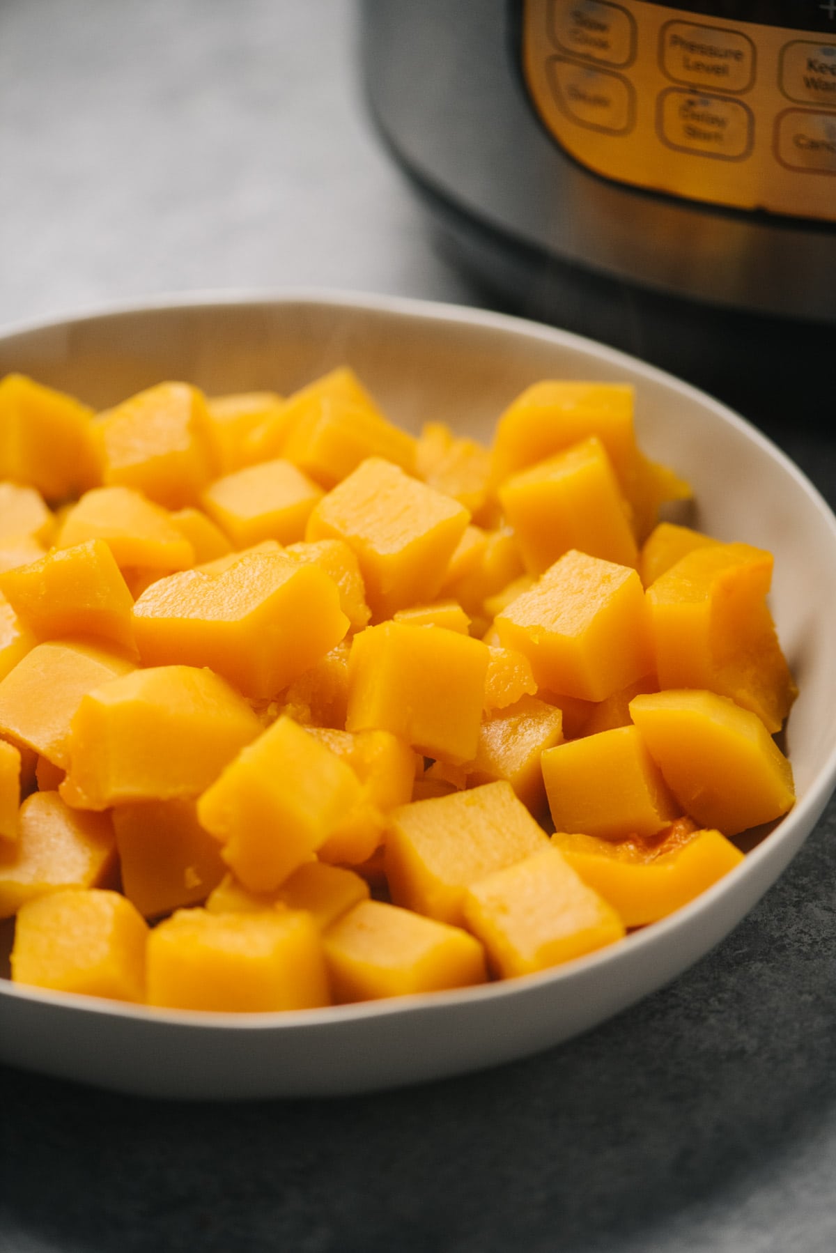 A bowl of steamed butternut squash cubes steamed in the instant pot on a concrete table with a pressure cooker in the background.