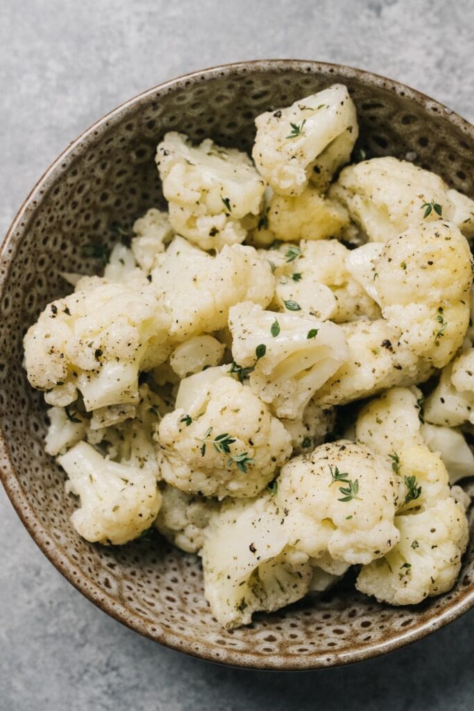 Steamed cauliflower florets tossed with olive oil, salt, and fresh herbs in a brown speckled bowl on a concrete table.