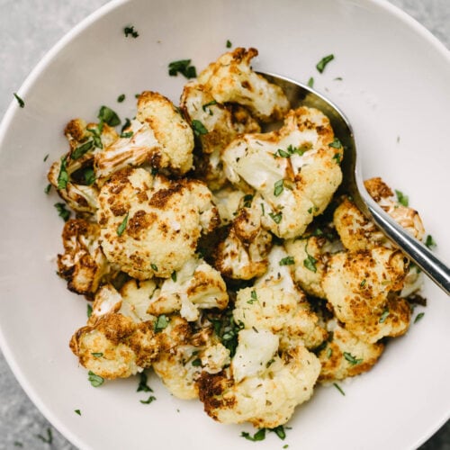 A spoon tucked into a bowl of roasted cauliflower florets, garnished with fresh chopped basil.