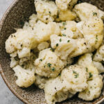 Steamed cauliflower florets tossed with olive oil, salt, and fresh herbs in a brown speckled bowl on a concrete table.