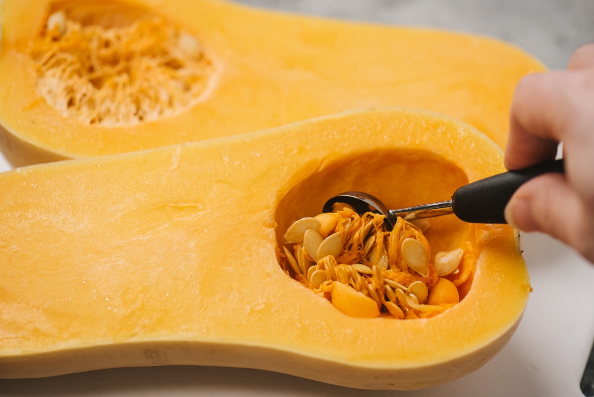 Using a melon baller to scoop seeds from the base of a butternut squash.