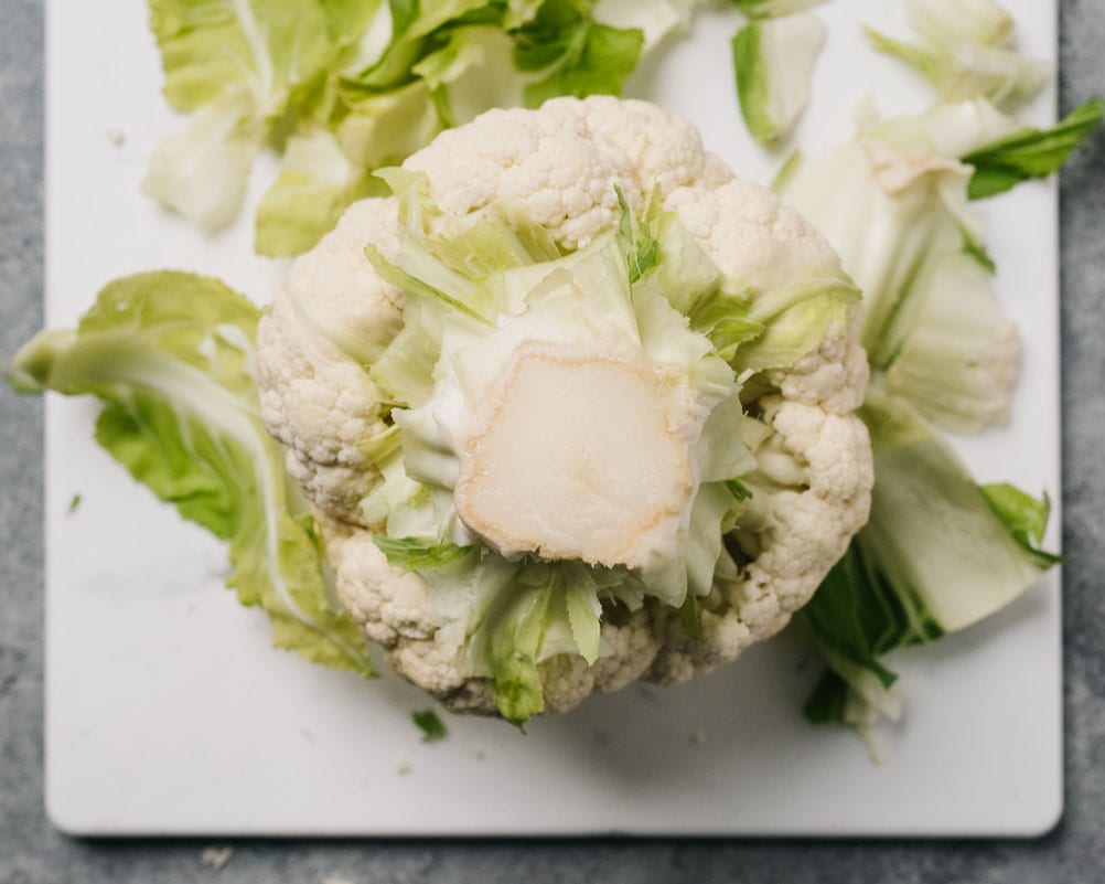 A head of cauliflower on a cutting board with all leaves remove and the stem fully exposed.