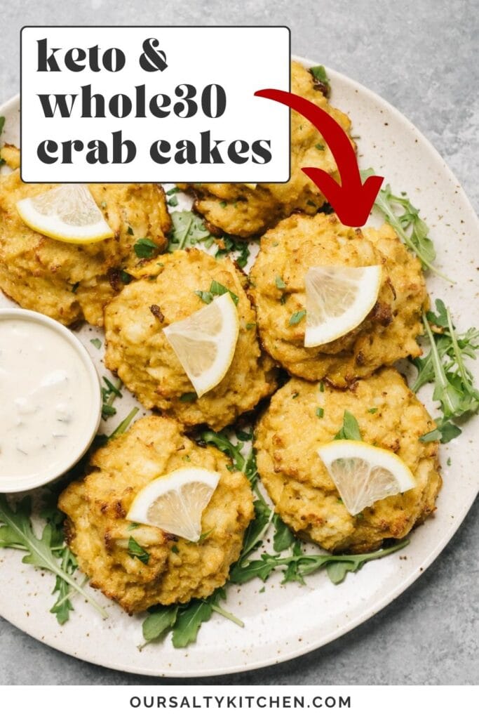 Six keto crab cakes on a speckled cream platter. A small dish of tartar sauce is on the plate, and each crab cake is garnished with a lemon slice; text overlay reads "keto and Whole30 crab cakes".