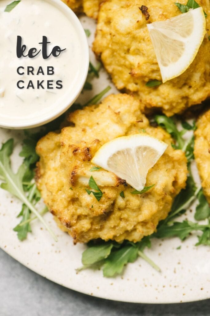 Several keto crab cakes on a cream speckled platter, garnished with lemon slices; small bowl of tarter sauce is to the side, and a text overlay reads "keto crab cakes".