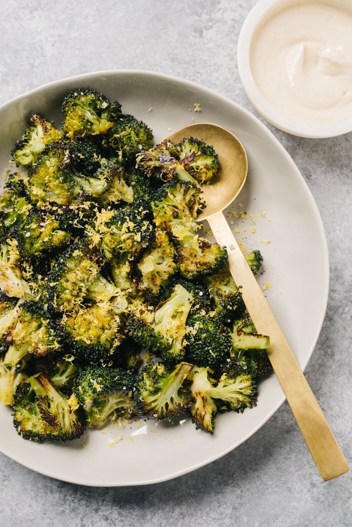 Roasted broccoli seasoned with lemon zest in a tan serving bowl with a gold serving spoon and small bowl of tahini sauce on the side.