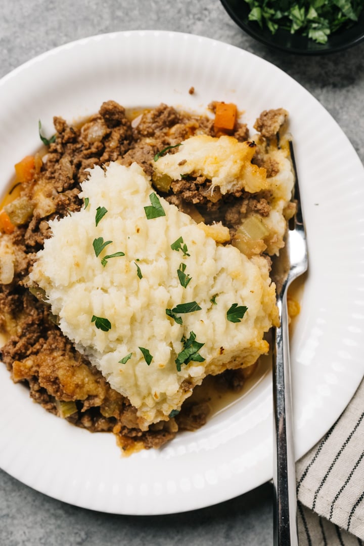 A serving of low carb shepherd's pie on a white plate with a silver fork on a concrete surface with a striped linen napkin.