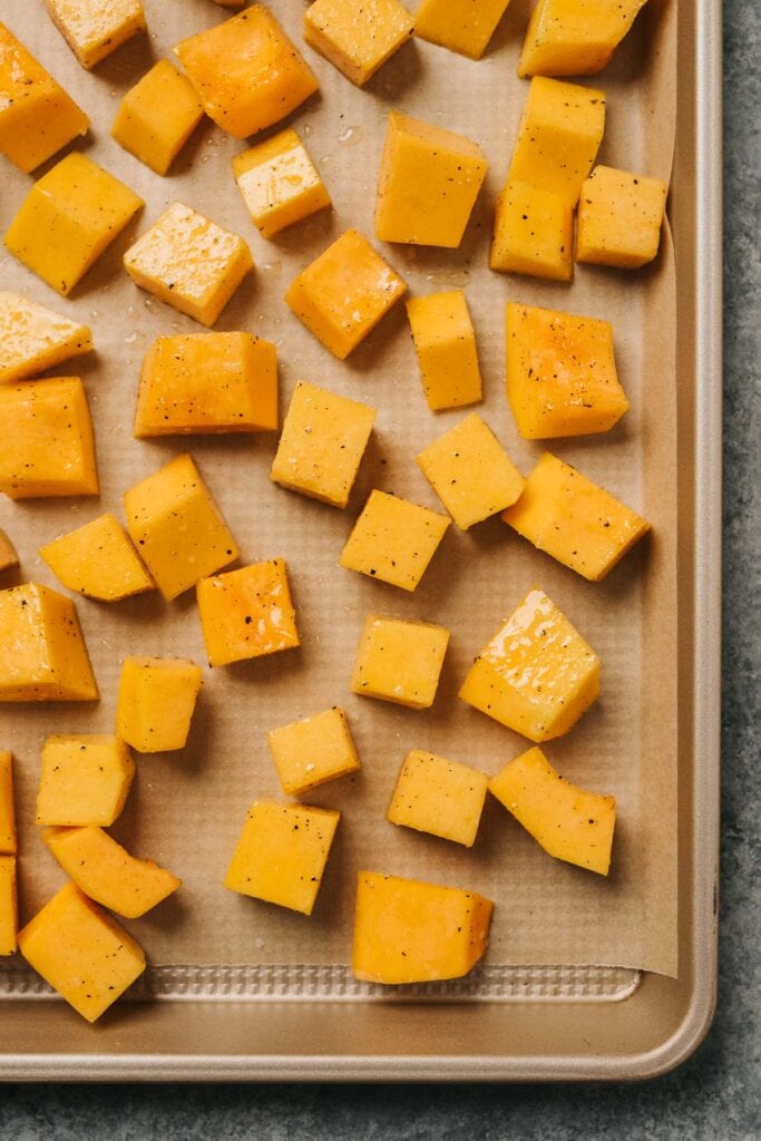 Diced butternut squash tossed with olive oil and seasonings arranged in an even layer on a parchment lined baking sheet.