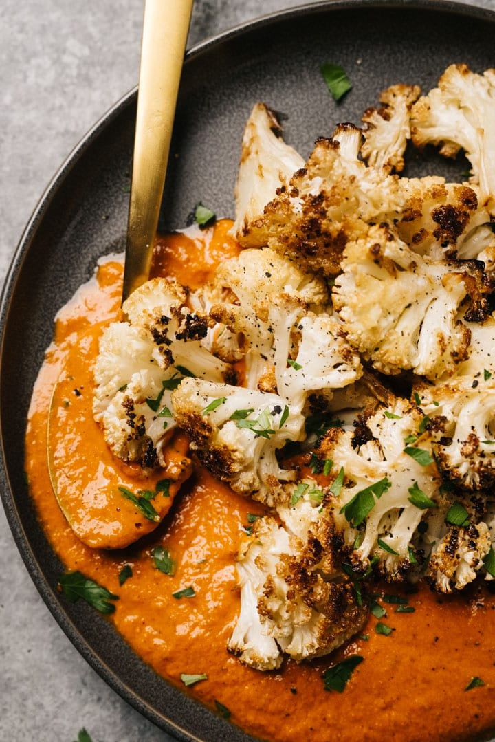 Side view, roasted cauliflower with romesco sauce on a grey plate with a gold serving spoon.