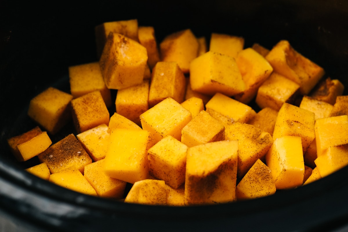 Cubed pieces of butternut squash in a slow cooker sprinkled with salt, cinnamon, and nutmeg.