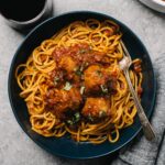 Spaghetti tossed with sunday sauce and topped with meatballs in a blue pasta bowl on a cement table with a glass of red wine, small bowl of chopped basil, and wedge of parmesan cheese.