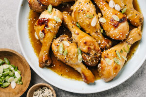 Baked chicken drumsticks with honey garlic sauce on a blue platter with small garnish bowls of sliced green onions and sesame seeds.