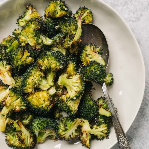 Oven roasted broccoli in a tan serving bowl with a silver serving spoon on a concrete background.