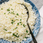 Cooked cauliflower rice in a blue speckled bowl with a silver spoon and a tan linen napkin.