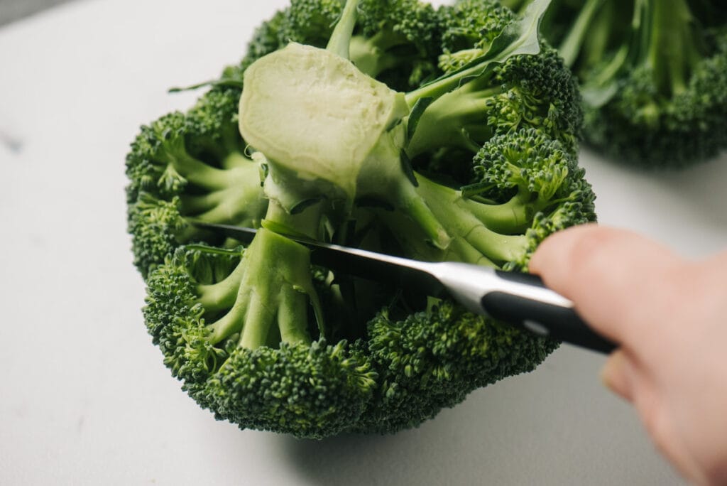 A paring knife cutting one floret away from a head of broccoli.