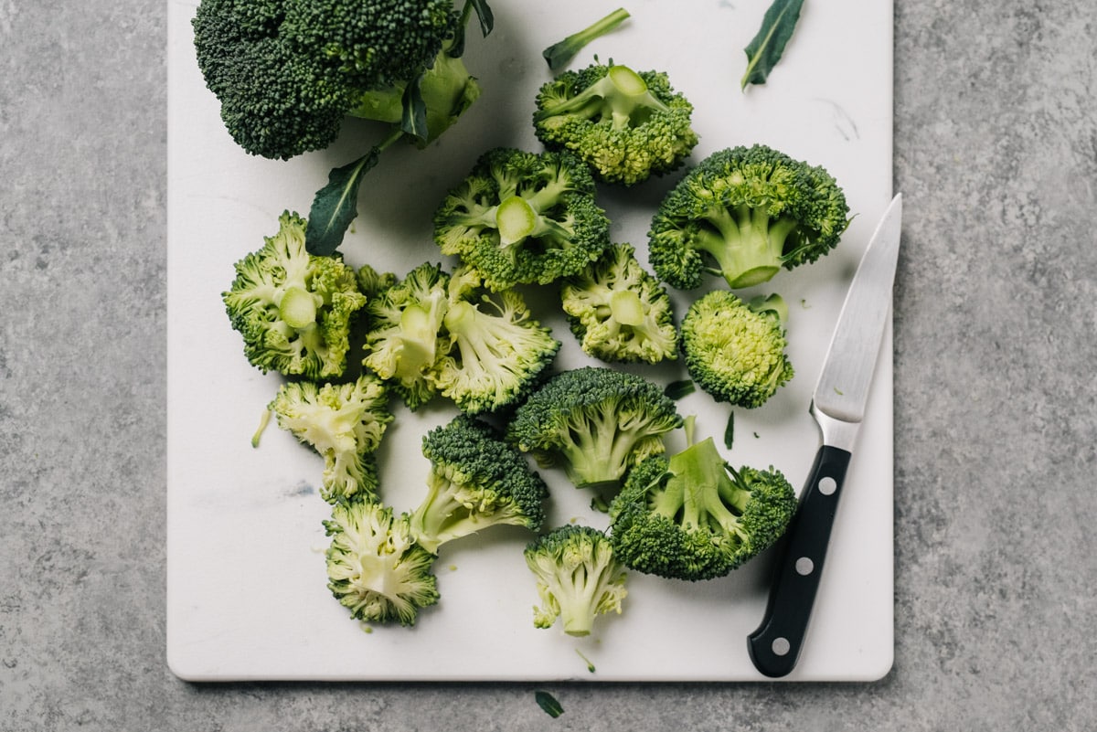 A head of broccoli trimmed down to florets on a cutting board with a paring knife.
