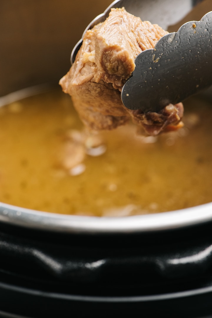 Tongs removing a chunk of cooked carnitas from the inner pot of an instant pot.
