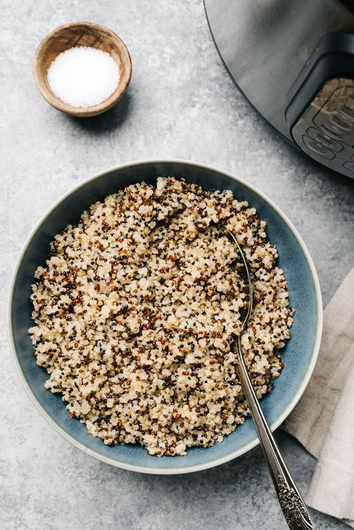 A blue bowl of quinoa on a concrete background with an instant pot and tan linen napkin to the side.
