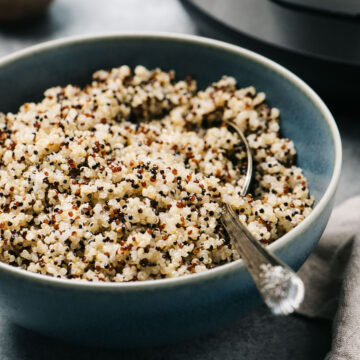 Side view, instant pot quinoa in a blue serving bowl with a silver serving spoon with an instant pot in the background and tan linen napkin to the side.