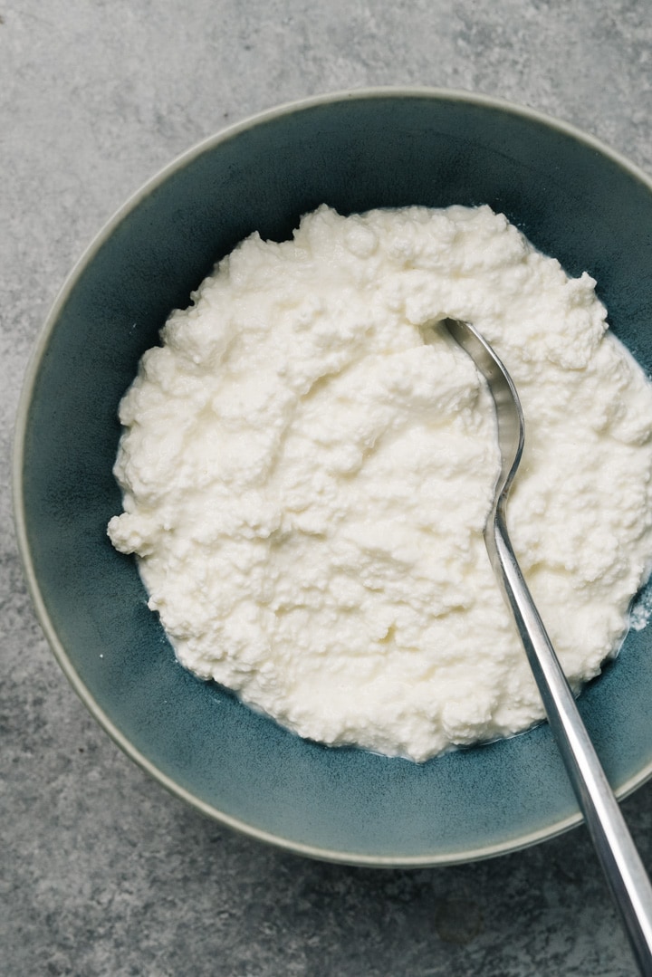 A blue bowl of homemade ricotta cheese with a silver spoon.