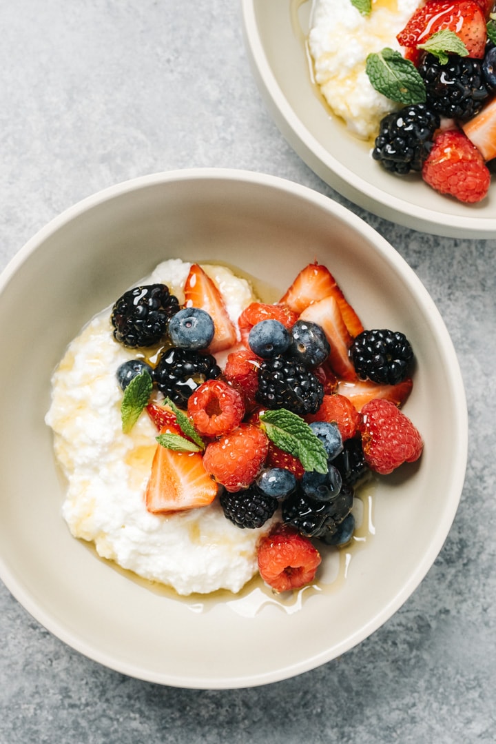Two bowls of fresh homemade ricotta cheese topped with berries, honey, and mint leaves.