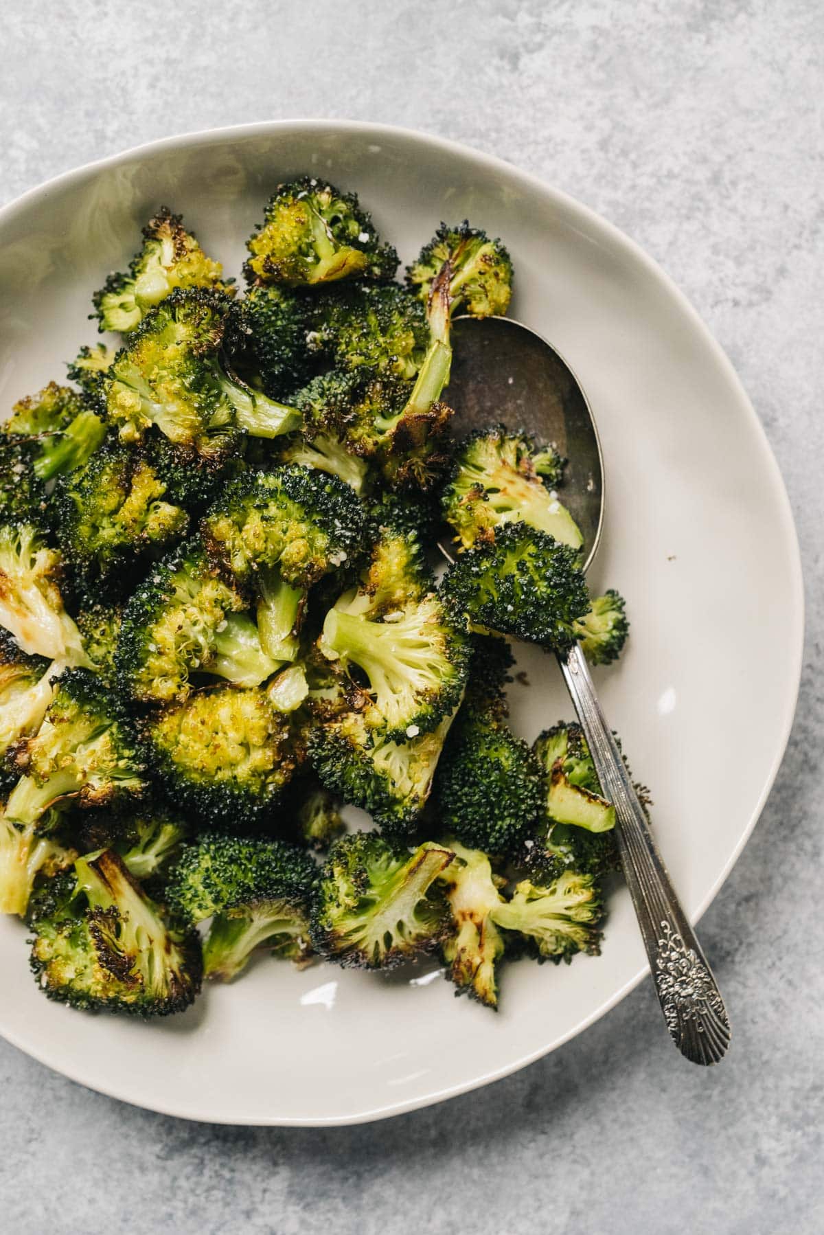 A bowl of roasted broccoli with a silver serving spoon on a concrete background.