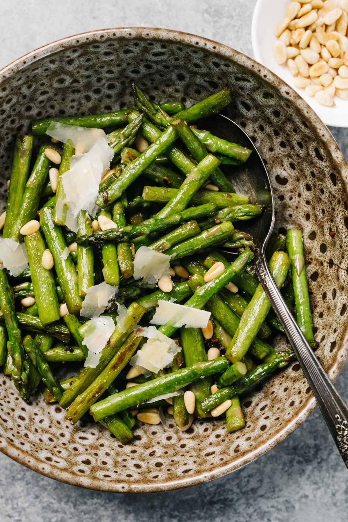 Sautéed asparagus in a brown speckled bowl with toasted pine nuts and shaved parmesan cheese.