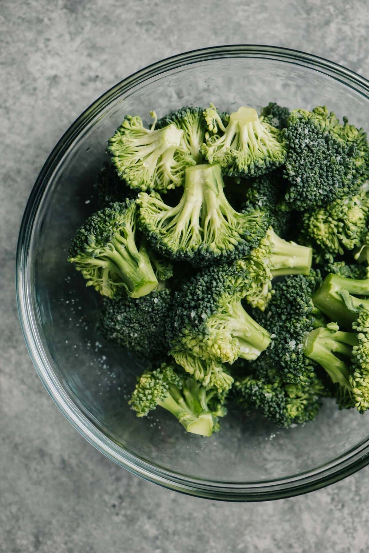 A bowl of raw broccoli florets sprinkled with salt with water in a glass microwave safe bowl.