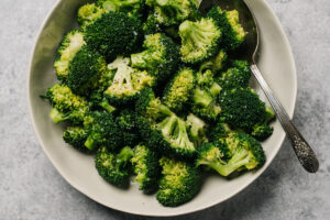 Steamed broccoli florets tossed with butter in a tan serving bowl with a silver serving spoon.
