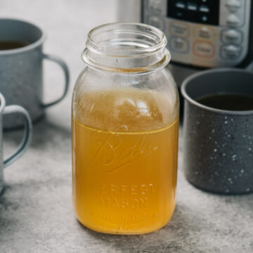 A mason jar of bone broth and 3 mugs of bone broth in front of an instant pot.
