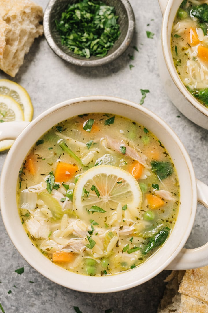 A bowl of lemon chicken orzo soup garnished with a lemon wheel and fresh parsley on a concrete background surrounded by a bowl of chopped parsley, lemon slices, and crusty bread.