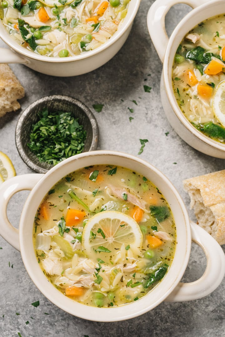 Several bowls of lemon chicken orzo soup garnished with a lemon wheel and fresh parsley on a concrete background surrounded by a bowl of chopped parsley, lemon slices, and crusty bread.