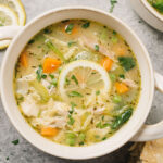 A bowl of lemon chicken orzo soup garnished with a lemon wheel and fresh parsley on a concrete background surrounded by a bowl of chopped parsley, lemon slices, and crusty bread.