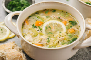 Side view, a bowl of lemon chicken orzo soup garnished with fresh parsley and a lemon wheel.