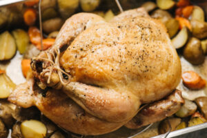 A whole roasted chicken in a roasting pan surrounded by carrots and potatoes.
