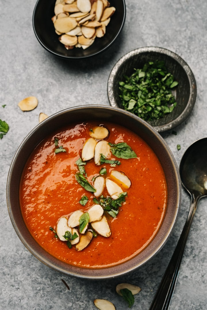 A bowl of roasted red pepper and tomato soup with small pinch bowls of basil and sliced almonds and a soup spoon on a concrete background.