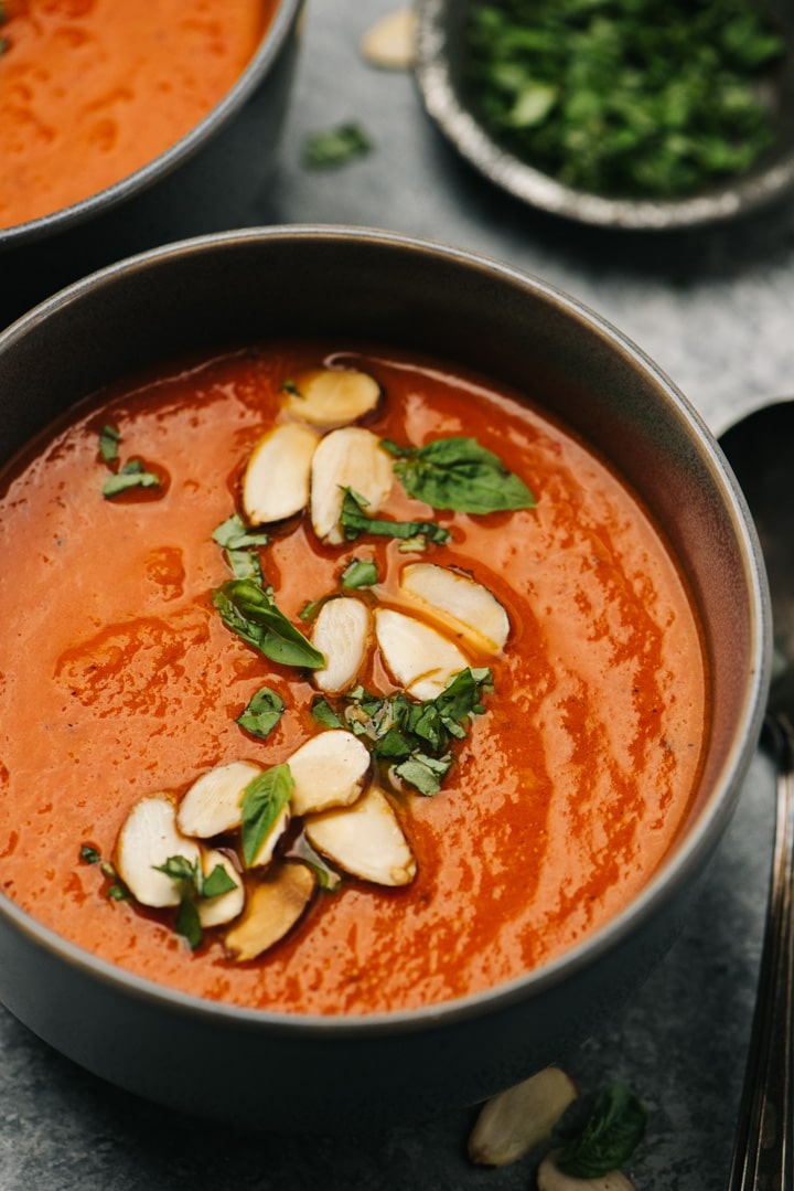 Side view, a bowl of roasted red pepper soup garnished with almonds and basil, with a small garnish bowl of basil in the background.
