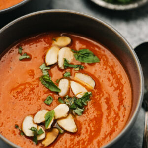Side view, a bowl of roasted red pepper soup garnished with almonds and basil.