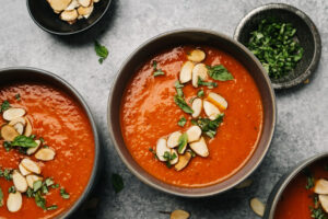 Several bowls of roasted red pepper and tomato soup with small pinch bowls of basil and sliced almonds and a soup spoon on a concrete background.