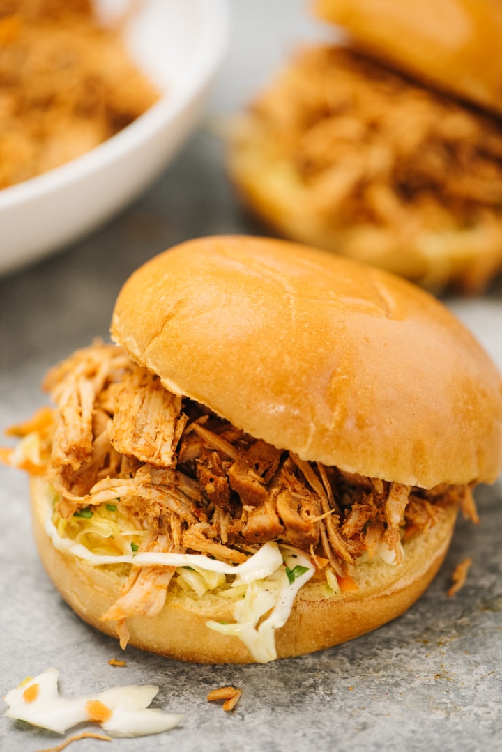 Two pulled pork sandwiches with coleslaw on a grey background, with a bowl of slow cooker pulled pork in the background.