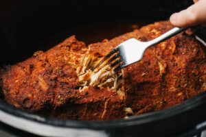 A fork pulling shreds of pulled pork in a slow cooker.