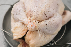 Side view, a trussed chicken seasoned with salt and pepper on a trivet on a grey plate.