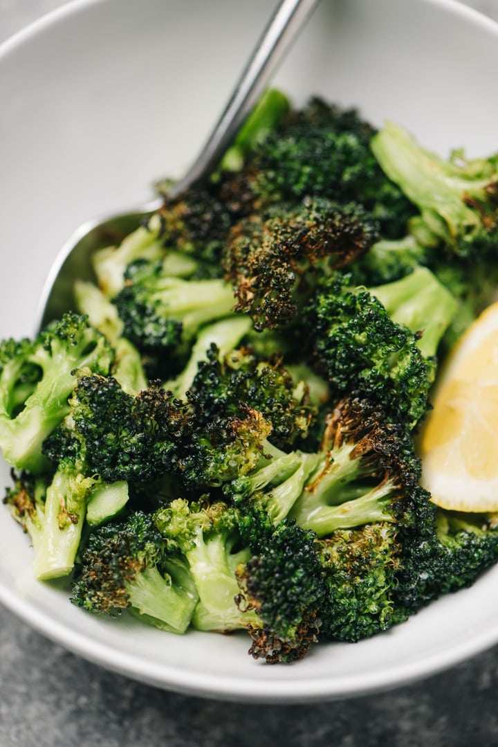 Side view, air fryer broccoli in a white bowl with a lemon wedge and silver serving spoon.