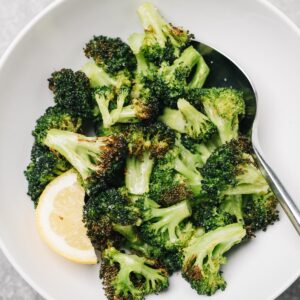 Air fryer broccoli florets in a white serving bowl with a lemon wedge on a concrete background.